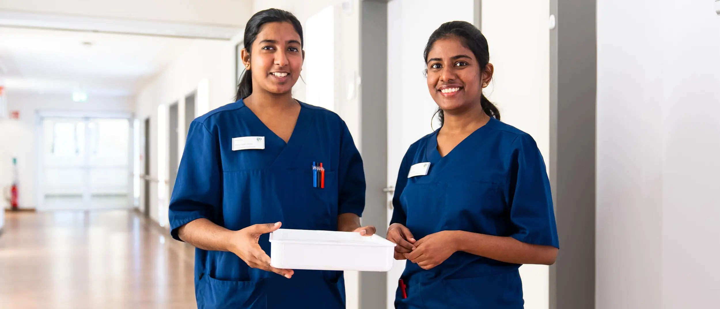 Two nurses in blue uniforms stand in the hospital corridor holding a white box.
