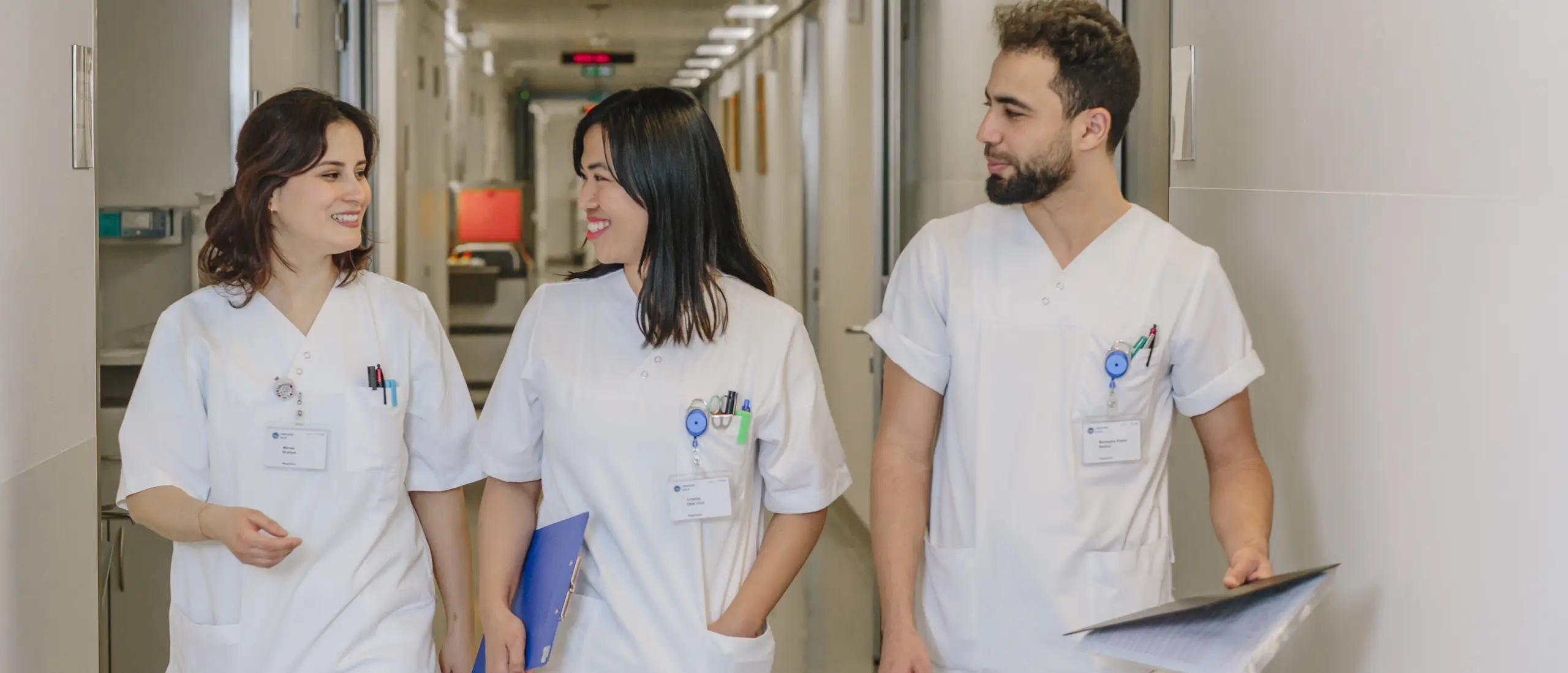 Three nurses in white work uniforms walk down a hospital corridor smiling.