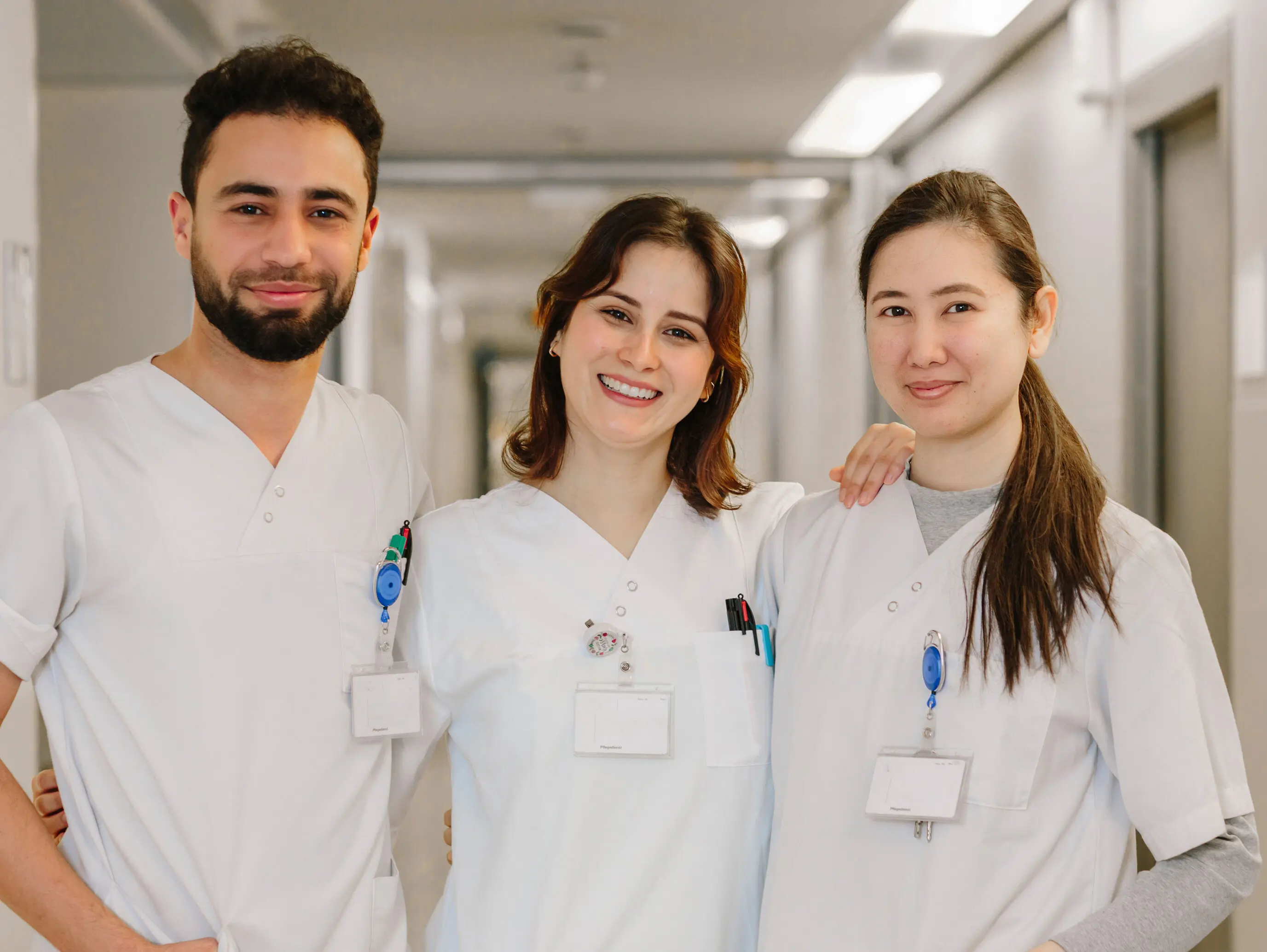 Three cheerful nurses in white work uniforms stand in a hospital corridor and smile at the camera.