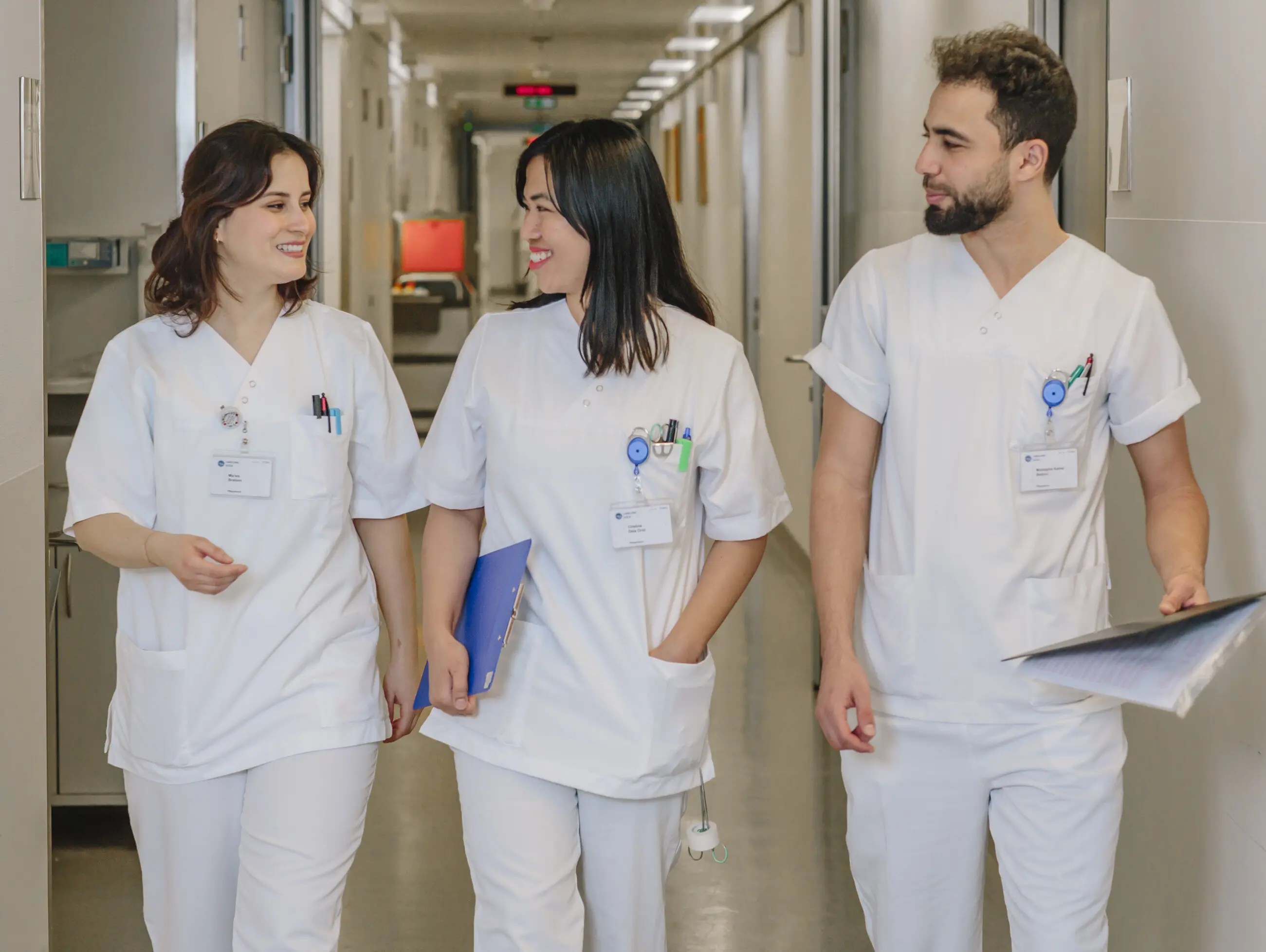 Three nurses in white work uniforms walk down a hospital corridor smiling.