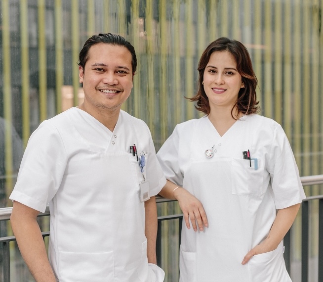 Two medical professionals in white stand smiling in front of a modern background.