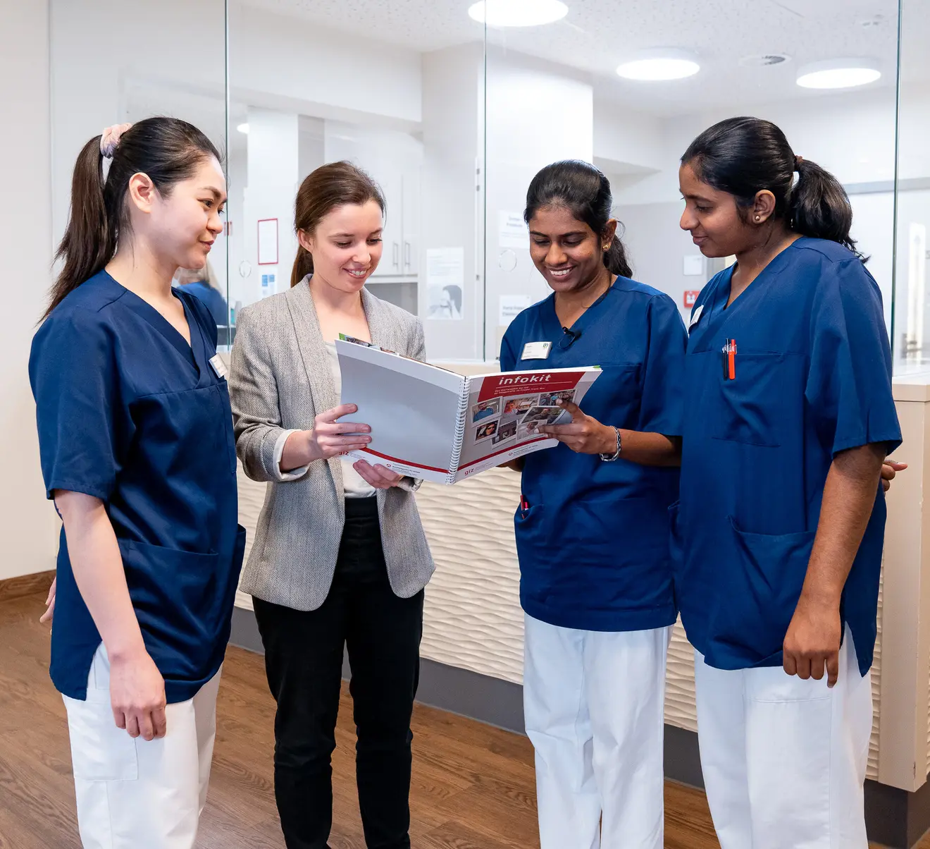 Dans un environnement m&eacute;dical moderne, quatre personnes consultent ensemble un livre. Trois d'entre elles portent des v&ecirc;tements de travail bleus, tandis qu'une personne est v&ecirc;tue d'un blazer gris.