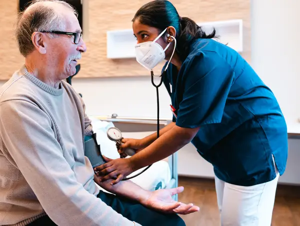 Un homme &acirc;g&eacute; est assis sur une table d'h&ocirc;pital tandis qu'une infirmi&egrave;re prend sa tension. Tous deux portent des masques et l'environnement est clair et moderne.