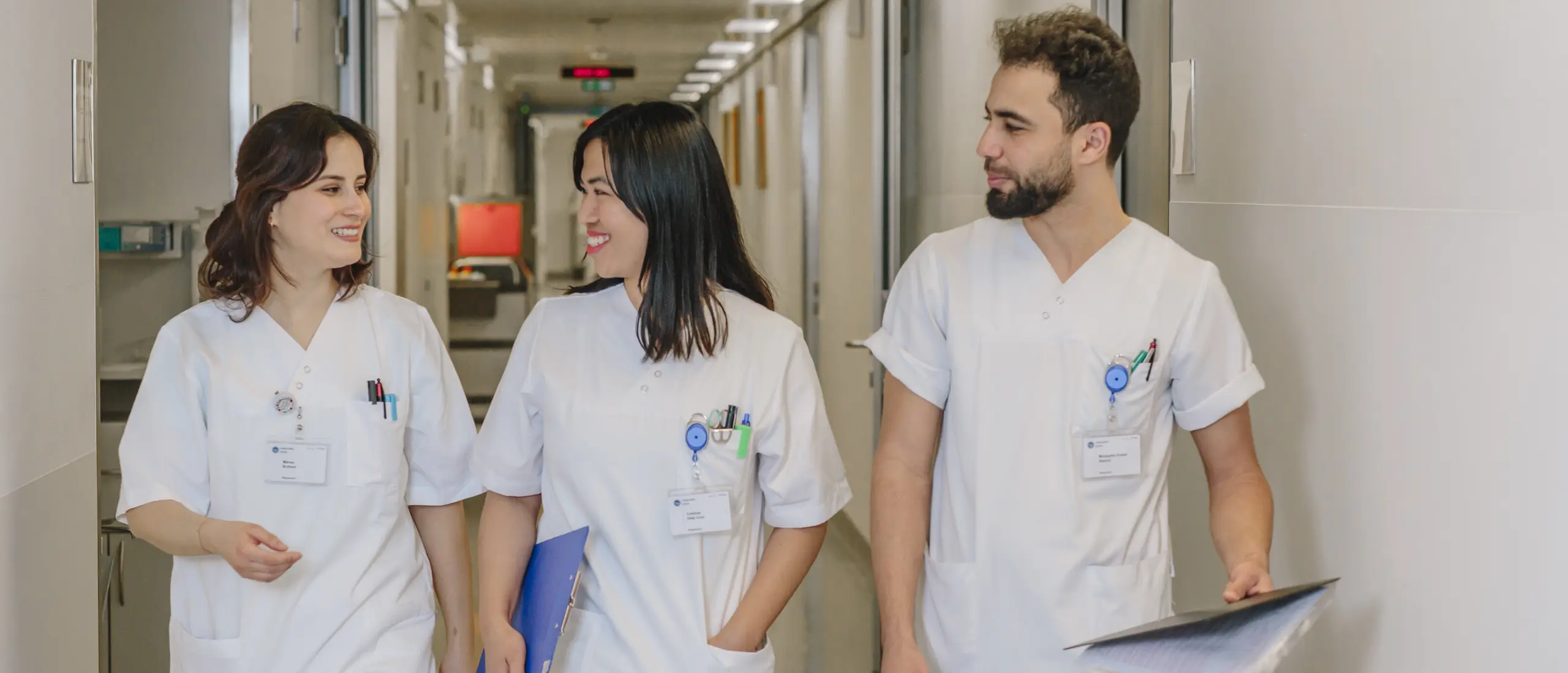 Trois m&eacute;decins en blouse blanche traversent un couloir d'h&ocirc;pital en souriant.