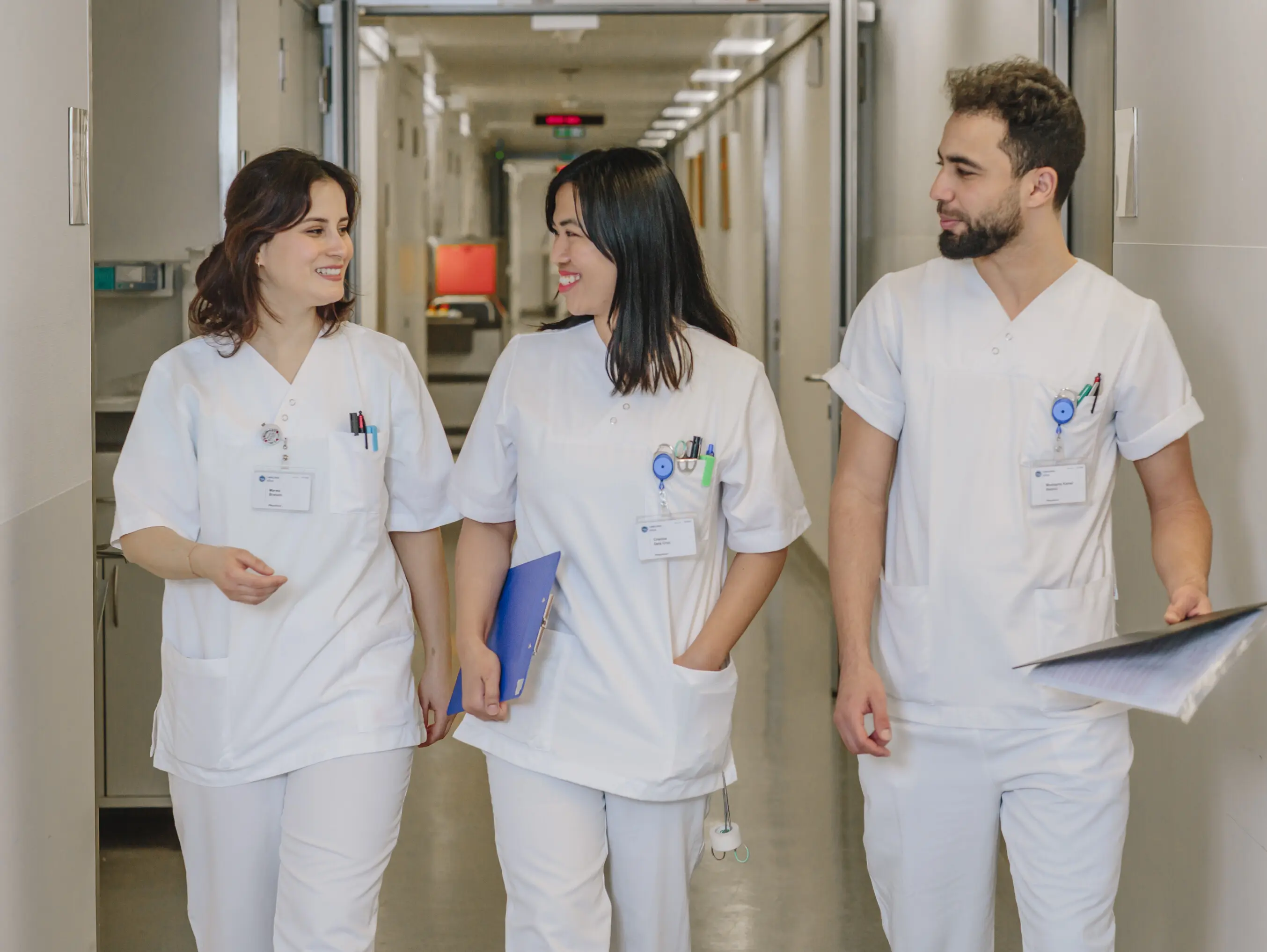 Trois m&eacute;decins en blouse blanche traversent un couloir d'h&ocirc;pital en souriant.