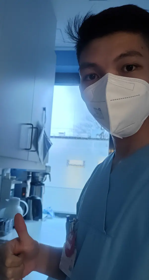 Un jeune homme en v&ecirc;tements de travail bleus et portant un masque m&eacute;dical, levant le pouce, se tient dans un environnement clinique lumineux.