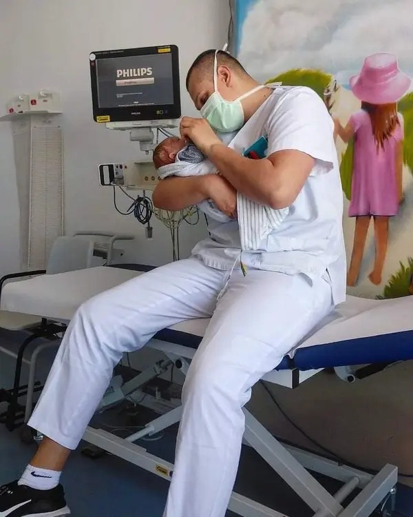 Dans la salle m&eacute;dicale, un infirmier tient un nouveau-n&eacute; dans ses bras et le regarde. Un &eacute;cran et une peinture murale sont visibles &agrave; l'arri&egrave;re-plan.